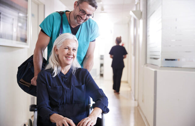 A male orderly pushing a woman in a wheelchair down a medical hall.