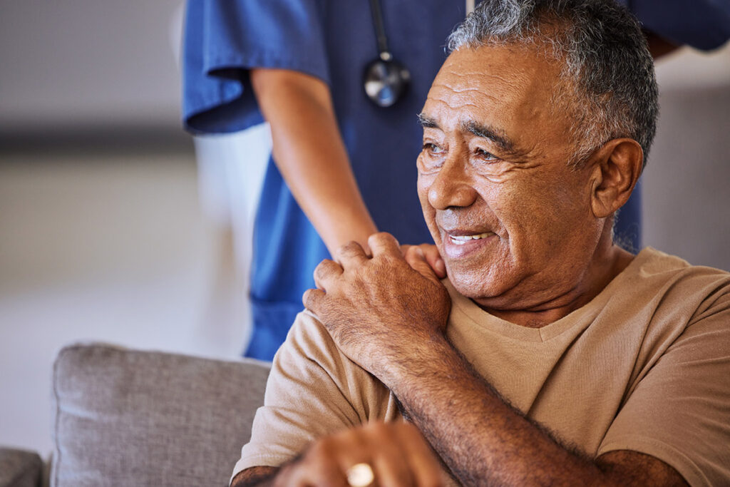 A nurse reassuringly touching the shoulder of her patient, who is smiling.